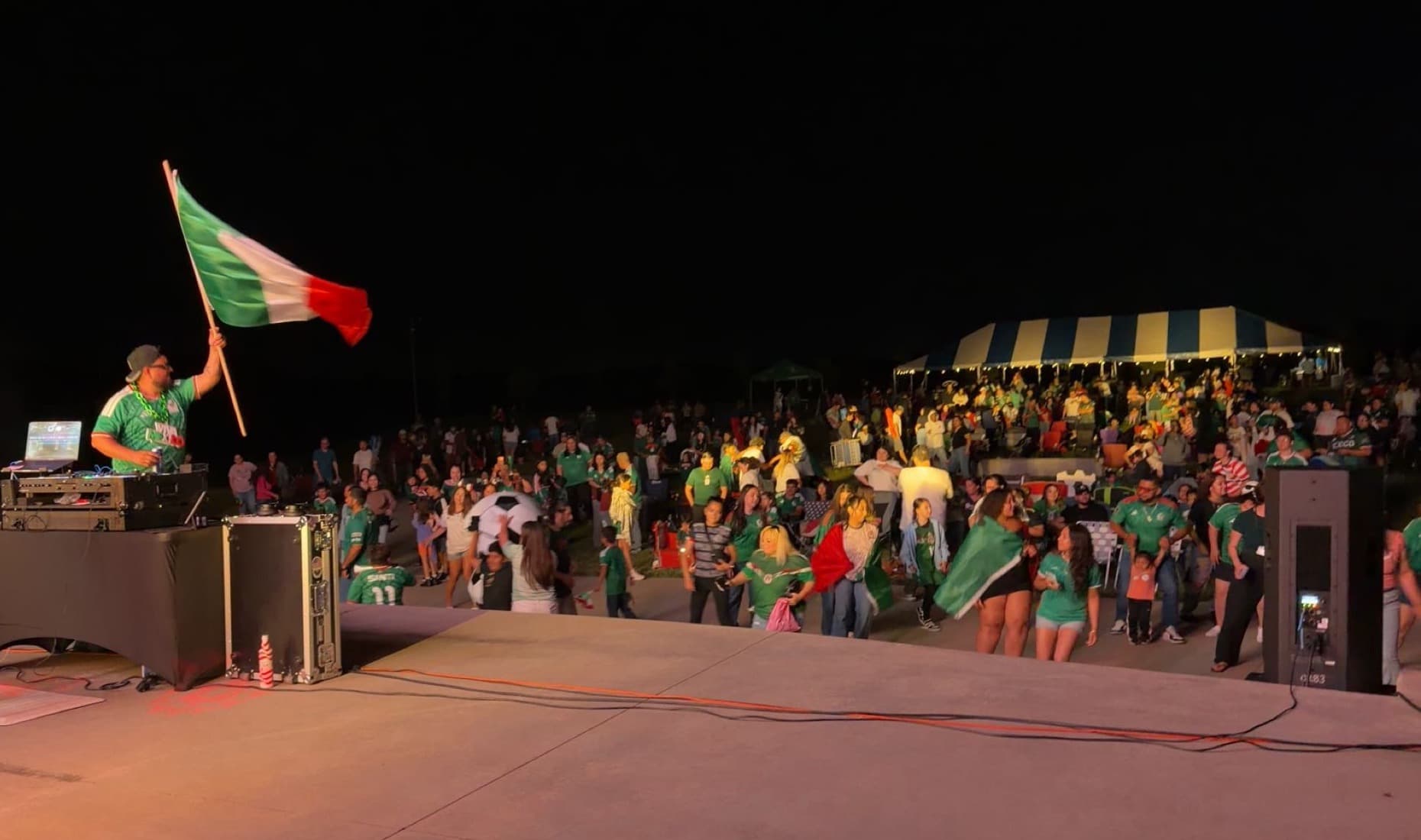 DJ TD waving a Mexican flag onstage while a crowd celebrates at an outdoor community event