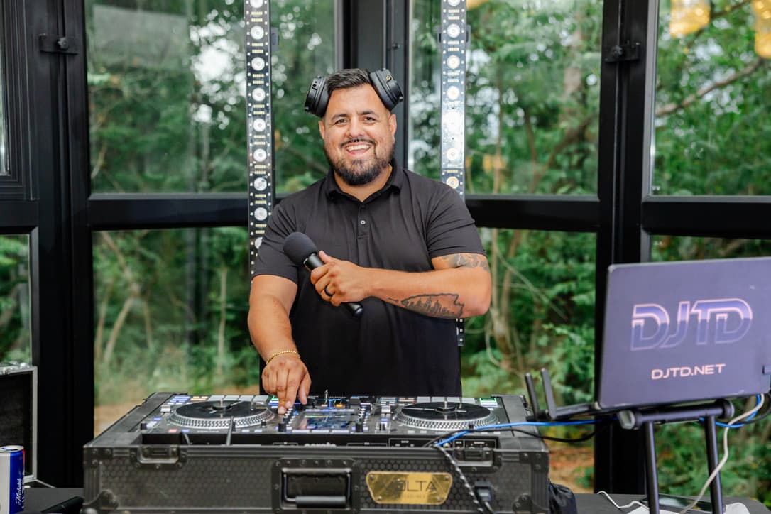 DJ TD smiling behind his DJ console, wearing headphones and holding a microphone.