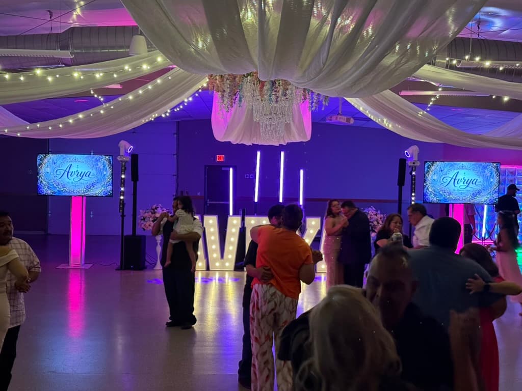 Guests dancing at a quinceañera beneath draped fabric and string lights, with purple lighting and personalized video displays.